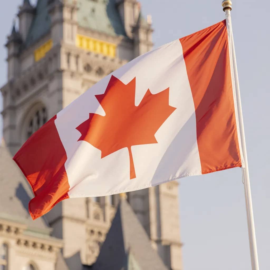 Le drapeau canadien avec une feuille d’érable rouge flotte au premier plan, avec un bâtiment historique orné d’une tour de l’horloge et d’un toit vert en arrière-plan flou.